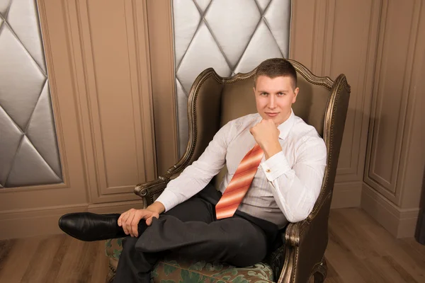 Confidence handsome businessman in chic interior. Successful young man in a business suit sitting on the armchair with his legs crossed and smiling — Stock Photo, Image