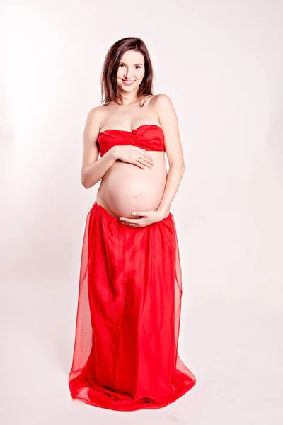 Красивая беременная девушка обнимает свой голый животик. красивая девушка в красной юбке и красный бюстгальтер стоя на белом фоне — стоковое фото