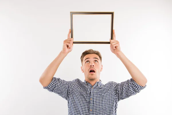 Hombre guapo sosteniendo un marco de imagen sobre su cabeza y mirando — Foto de Stock