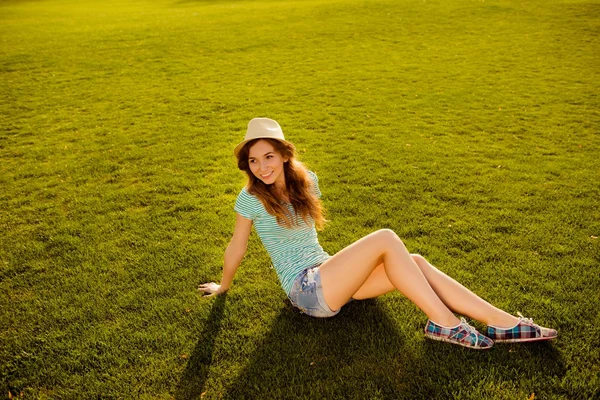Fröhliches junges Mädchen mit Hut sitzt auf dem Gras — Stockfoto