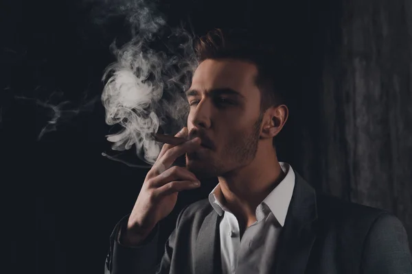 Handome confident man in suit on the grey background smoking a c — Stock Photo, Image