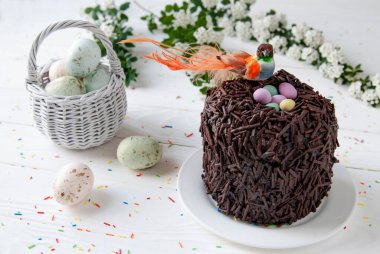 Paskalya çikolatalı keki ve yuvada oturan kuş. Küçük turuncu kuş, renkli yumurta ve hasır sepet Paskalya dekorunun arka planında