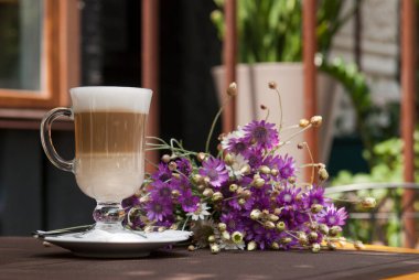 Restoranın yaz terasında bir bardak latte ve mor çiçekler.
