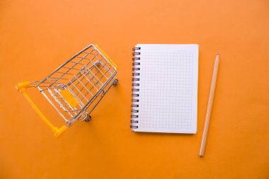 Üst manzara not defteri, alışveriş arabası ve kalem, turuncu arka planda izole edilmiş. Uzayı kopyala, düz yat.