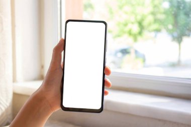 Kadın pencere kenarında oturuyor ve beyaz masaüstü ekranlı cep telefonu kullanıyor. Model telefon görüntüsü.