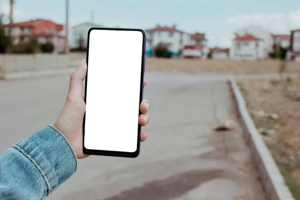 Mockup image of woman hand holding blank white desktop screen mobile phone. Blurry small city and street in the background. Female hand using smartphone with copy space for your advertise text.