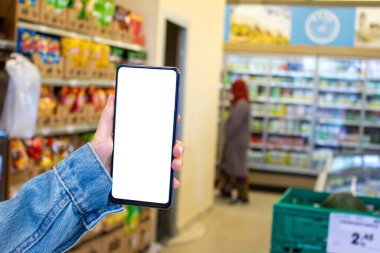 Süpermarkette elinde beyaz masaüstü cep telefonu olan bir kadının resmi..