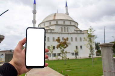 Beyaz masaüstü ekranlı cep telefonu tutan adamın görüntüsü. Arka planda bulanık cami görünümü.