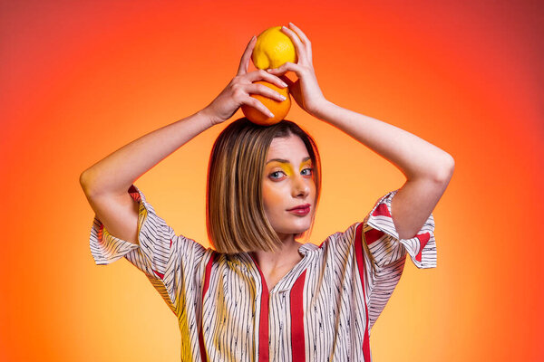 Beautiful attractive blond young woman light blue eyes holding orange on head with bright yellow background - Studio photography of pretty woman who holds citrus lemon on top