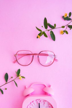 Resimde oval gözlükler var. Pastel pembe arka planda, yaz tarzında. Çalar saat, sarı çiçekler ve yeşil izin.