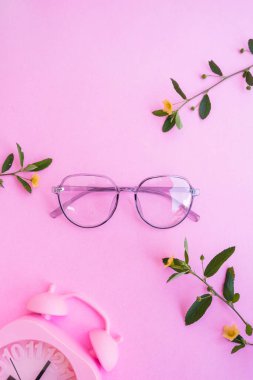 Resimde oval gözlükler var. Pastel pembe arka planda, yaz tarzında. Çalar saat, sarı çiçekler ve yeşil izin.