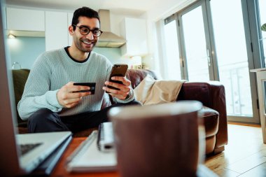 Karışık ırk işadamı koltukta oturmuş gülümsüyor internette ödemeler yapıyor cep telefonu tutuyor.