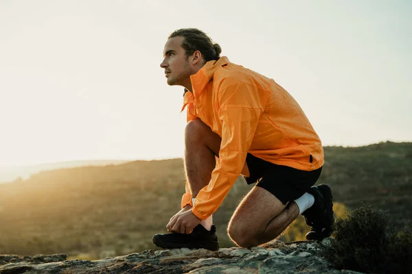 Atleta masculino caucásico tomando un descanso de correr atando cordones de zapatos mientras camina en la montaña durante el atardecer — Foto de Stock