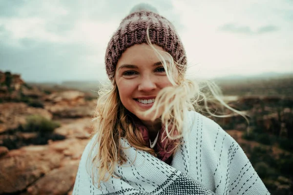 Donna caucasica sorridente che indossa berretto avvolto in accogliente coperta godendo di campeggio sulla cima della montagna — Foto Stock