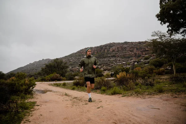 Hezký mladý samec sprinter trénink v oblačném počasí běží na štěrkové horské stezce se zelenými keři a kopci — Stock fotografie