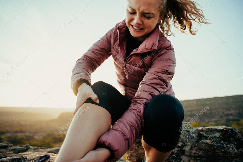 Close up injured female hiking holding sprained knee sitting on rock at sunset