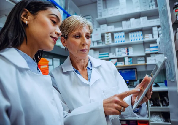 Elderly pharmacist guiding new female intern teaching scrolling on digital tablet standing in pharmacy