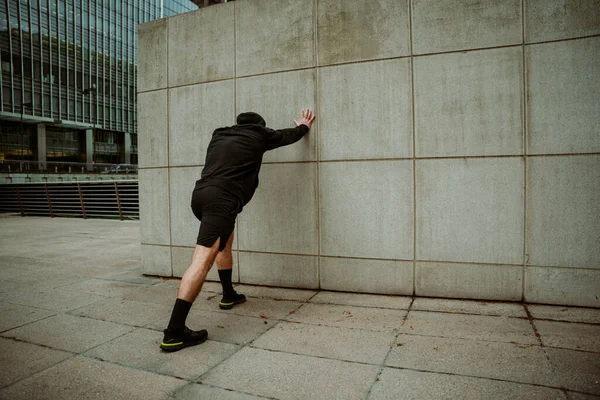 Caucasian male athlete stretching against wall after long run in city