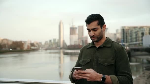 Mixed race male young adult typing on smartphone standing on bridge overlooking lake in the city — Stock Video