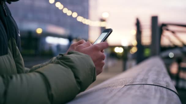 Close up gemischte Rasse männlichen jungen Erwachsenen SMS auf dem Smartphone lehnt an Geländer der Brücke in kalten Stadt Abend — Stockvideo