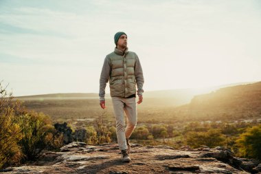 Beyaz tenli genç erkek, gün batımında sıcak giysiler içinde yabanda yürüyor.