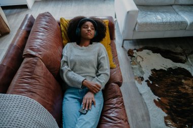 Kulaklık takan melankolik bir kadın modern salonda deri koltukta uzanmış akıllı telefonu tutarken hüzünlü hareketler dinliyor..