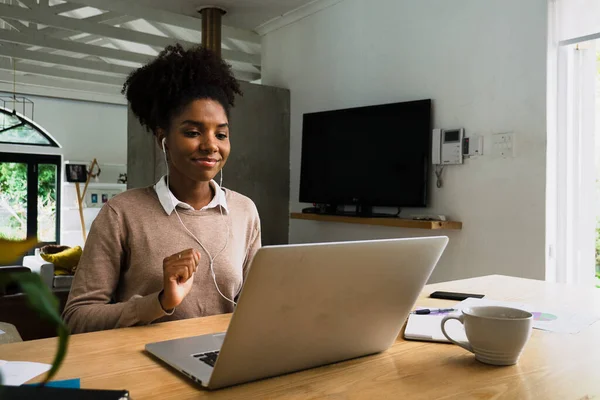 Sorridente donna africana con auricolari afro mentre si impegna in videochiamate con colleghi di lavoro. Seduto al tavolo della cucina con caffè caldo — Foto Stock