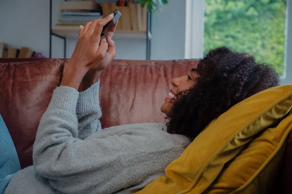 Mutlu melez kadın akıllı telefondan heyecan verici mesajlar alırken rahat bir şekilde kanepede dinleniyor..