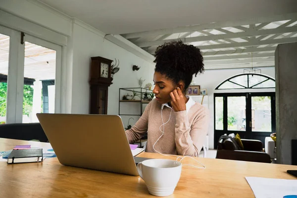 Occupata donna di lavoro mista che scrive scartoffie indossando auricolari che ascoltano videochiamate sul computer portatile seduto alla scrivania in cucina moderna. — Foto Stock