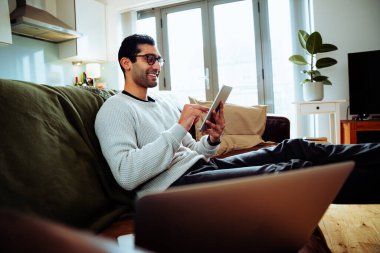 Karışık ırk işadamı evde çalışıyor koltukta oturup dijital tablette gülümsüyor. 