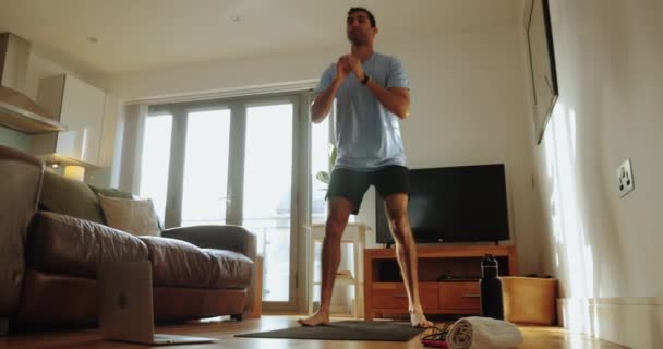 Masculino de raza mixta haciendo ejercicio en la sala de estar en cuclillas en la colchoneta del gimnasio sintiéndose bien antes del trabajo — Vídeos de Stock