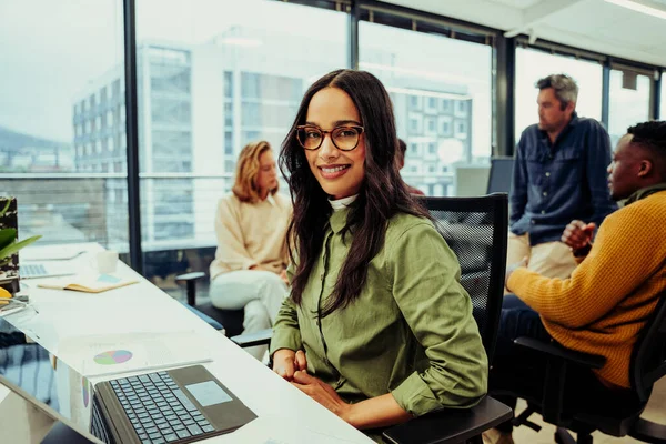 Bella designer indiana sorridente ricerca layout disegni sul computer portatile mentre il collega si siede dietro impegnarsi in una riunione di lavoro — Foto Stock