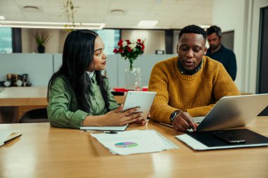 Dijital tabletle çalışan Hintli kadın, ofiste dizüstü bilgisayarda yazı yazan melez bir adamla çalışmayı tartışıyor.