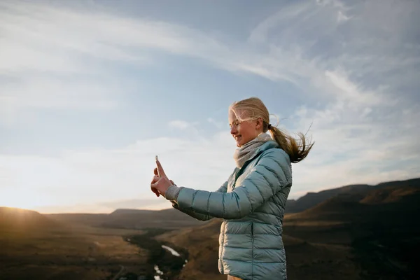 Young caucasian blonde female taking selfie on top of mountain while hiking at sunset. — Stock Photo, Image