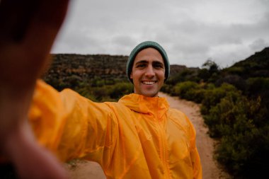 Çekici genç erkek sporcu, çalılarla dolu bulutlu bir havada çakıllı dağ yolunda koşarken kendi fotoğrafını çekerken gülümsüyor.