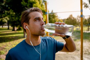 Susamış genç erkek sporcu kulaklıkla dışarıda egzersiz yaparken yukarı bakarak müzik dinliyor.