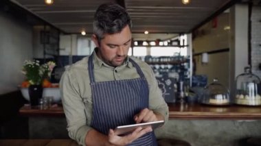 Yakışıklı kafe sahibinin tableti kaydırırken modaya uygun kahve dükkanının tezgahına yaslanması..