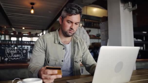 Jóképű kaukázusi férfi kezében hitelkártya fizetési laptop ül mellett forró kávé trendi kávézó. — Stock videók