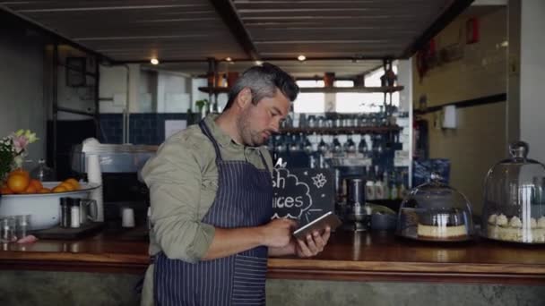 Beau propriétaire de café masculin portant tablier, se concentrant tout en faisant défiler sur la tablette, debout dans un café funky. — Video