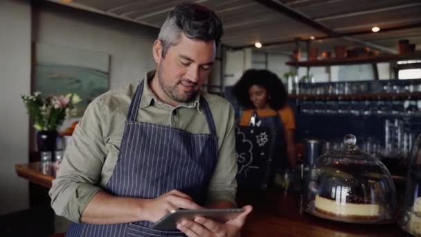 Clip de dueño de café guapo desplazándose en la tableta mientras hermosa hembra hacer café. — Vídeo de stock