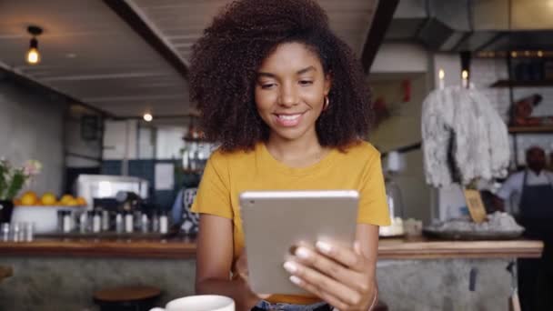 Piękna uśmiechnięta kobieta siedzi w modnej kawiarni przewijając na tablecie obok filiżanki gorącej kawy. — Wideo stockowe