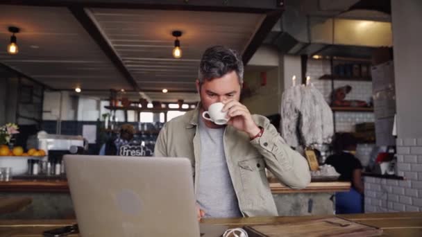 Όμορφος επιχειρηματίας πληκτρολογώντας στο laptop, ενώ πίνοντας ζεστό καφέ κάθεται σε funky cafe. — Αρχείο Βίντεο