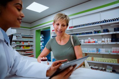 Kafkasyalı anne eczanedeki dijital tablete bakarak genç stajyer kimyagerle sohbet ediyor. 