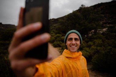 Yakışıklı, genç bir koşucu, gülümsüyor. Soğuk bulutlu bir havada dağ yolunda kendini akıllı telefondan çekiyor. 