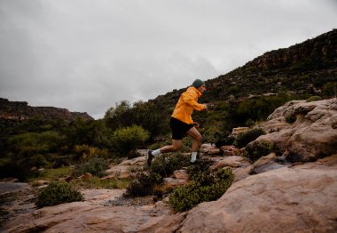 Güçlü erkek koşucu, bulutlu havada, yeşil çalılar ve dik tepelerle ıslak dağ kayalarını hızla koşuyor.