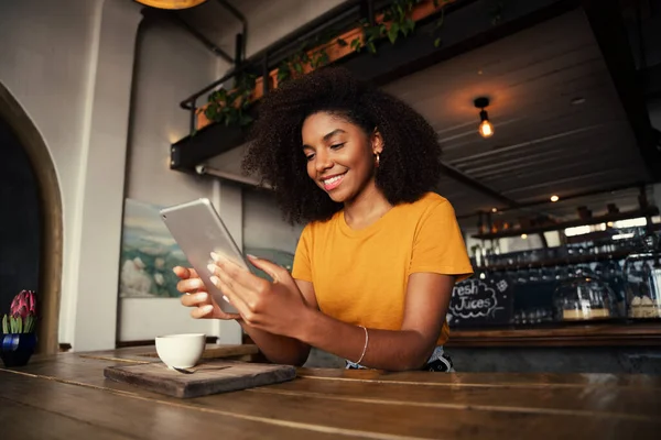 Basso angolo colpo di bella femmina afro-americana in possesso di tablet digitale seduto in sala da tè funky con latte caldo. — Foto Stock