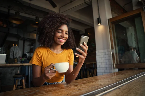 Bella donna afroamericana che scorre sullo smartphone alla guida di caffè in caffetteria alla moda. — Foto Stock