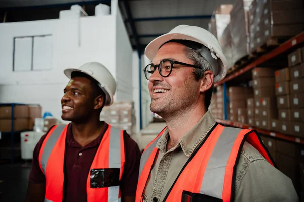 caucasian male worker wearing spectacles laughing with mixed race worker standing in factory warehouse.