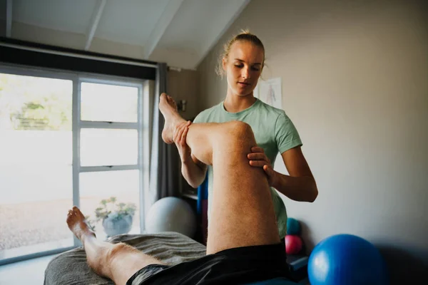 Vrouwelijke fysiotherapeut buigen knie naar borst van mannelijke patiënt liggend in pijn op massage bed in pilates kamer. — Stockfoto
