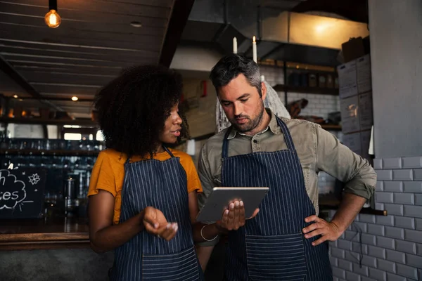 Colleghi curiosi che indossano grembiuli, discutono di finanze mentre guardano tablet in piedi in caffetteria. — Foto Stock